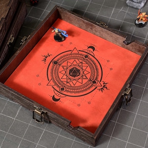 May include: A wooden dice tray with a red interior featuring a black geometric design. The tray is open, revealing the interior, and has metal clasps. Miniature figurines and dice are visible nearby.