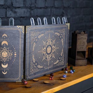 Op de afbeelding: Een donkere houten tabletop RPG-gameset. De set bevat een vouwscherm met hemelse ontwerpen, dobbelsteentorens, dobbelstenen en houten tokens. Het scherm heeft gouden accenten en doorzichtige markeringen.