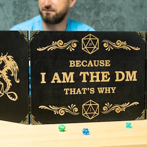 May include: A black wooden three-panel dungeon master screen featuring gold-coloured engraved dragon and twenty-sided die illustrations. The centre panel displays the text "BECAUSE I AM THE DM THAT'S WHY" in gold lettering with ornate scrollwork accents.