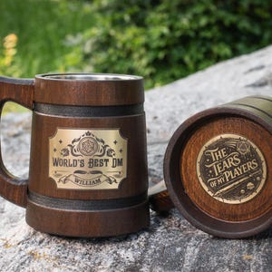 Puede incluir: Una jarra de madera marrón oscuro con interior de metal y asa. Una placa dorada en el frente dice "WORLD'S BEST DM" y "WILLIAM". Un posavasos de madera a juego presenta una incrustación circular dorada con el texto "THE TEARS OF MY PLAYERS".