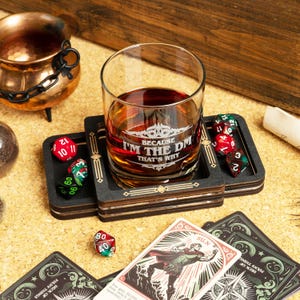 Puede incluir: Un vaso de whisky lleno de líquido ámbar descansa sobre un posavasos negro con detalles dorados. El vaso dice "BECAUSE I'M THE DM THAT'S WHY". Dados y cartas de juego están dispersos alrededor.