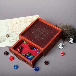 May include: A wooden dice box with a burgundy interior and a drawer filled with various colored dice. The box has a decorative geometric design on the lid. A map and miniature figurines are also present.