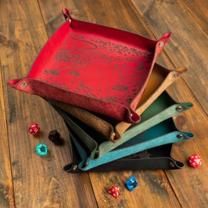 May include: A stack of colorful leather dice trays in red, burgundy, brown, green, teal, and black. Each tray has a snap closure and a map design with the text "Roll for Adventure". Several dice are scattered around the trays on a wooden surface.