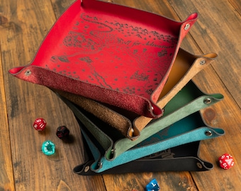 Könnte beinhalten: Ein Stapel farbiger Würfelablagen aus Leder in Rot, Burgunder, Braun, Grün, Türkis und Schwarz. Jede Ablage hat einen Druckknopfverschluss und ein Kartendesign mit dem Text "Roll for Adventure". Mehrere Würfel liegen auf einer Holzoberfläche verstreut.