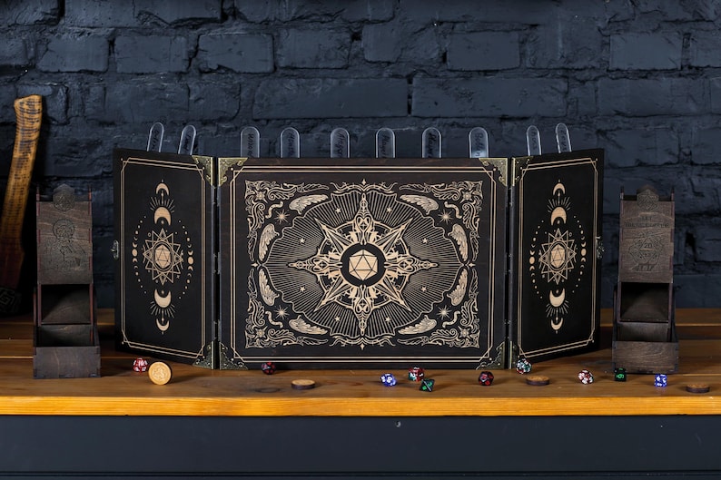 Op de afbeelding: Een zwart en goud tabletop rollenspel scherm met een gedetailleerd hemels ontwerp. Inclusief twee houten dobbelstenen torens, dobbelstenen en een munt op een houten oppervlak. Het scherm heeft doorzichtige plastic clips aan de bovenkant.
