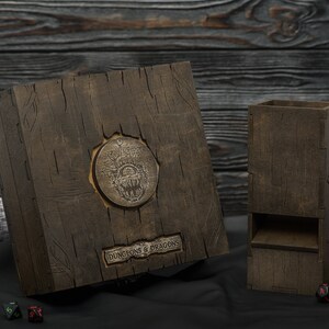 May include: A dark brown wooden dice box and dice tower for Dungeons & Dragons. The box features a metal emblem and the words "DUNGEONS & DRAGONS". The dice tower is also brown. Dice and figurines are visible.