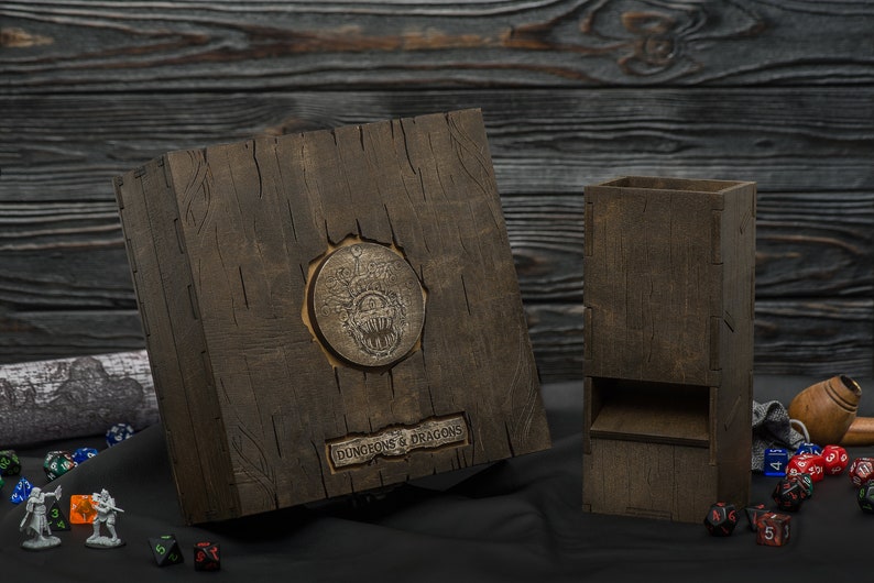 May include: A dark brown wooden dice box and dice tower for Dungeons & Dragons. The box features a metal emblem and the words "DUNGEONS & DRAGONS". The dice tower is also brown. Dice and figurines are visible.