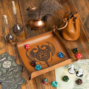 May include: A brown leather dice tray with a dragon and sword design, surrounded by various colorful dice, a leather pouch, and a deck of cards. The tray is on a wooden surface, with a copper pot and glass vials.