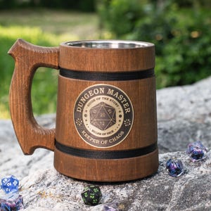 May include: A brown wooden tankard with a metal interior lining and a sturdy handle. The exterior features a circular emblem with the text "DUNGEON MASTER," "GUIDE OF THE STORY," and "KEEPER OF CHAOS" surrounding a twenty-sided die graphic.