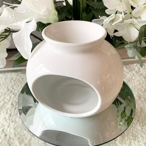 May include: White ceramic wax melt burner with a rounded shape and a large opening on the side. The burner is sitting on a glass surface.