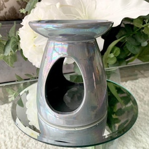 May include: A grey ceramic oil burner with an iridescent finish. The burner has a round top and a wide, oval-shaped opening in the centre. The burner is sitting on a round, mirrored surface.