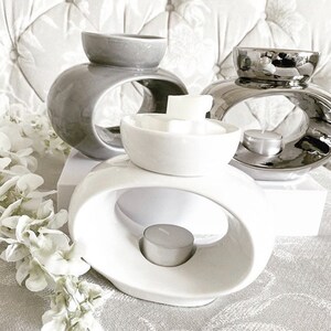 May include: Three ceramic wax melt warmers in grey, silver, and white. The warmers have a unique oval design with a ring in the centre to hold a tea light candle. The white warmer has a small bowl on top to hold wax melts.