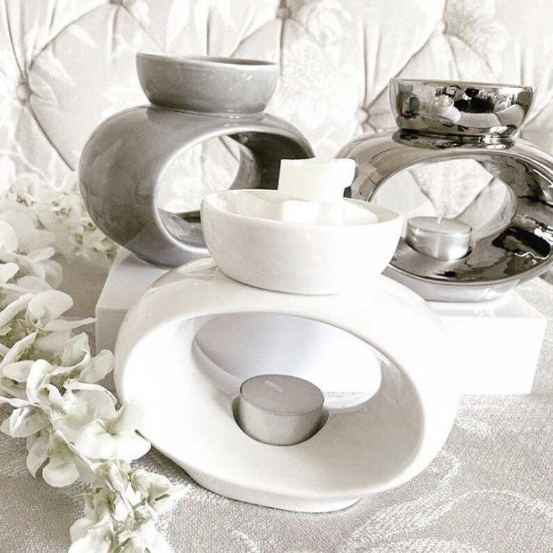 May include: Three ceramic wax melt warmers in grey, silver, and white. The warmers have a unique oval design with a ring in the centre to hold a tea light candle. The white warmer has a small bowl on top to hold wax melts.