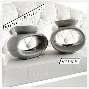 May include: Two grey ceramic oil diffusers with a unique oval design. The diffusers are sitting on a grey book with the word "Dior" printed on the cover.