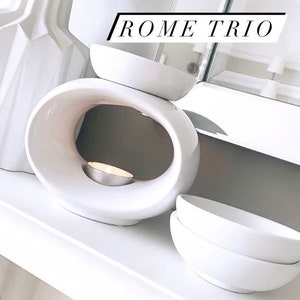 May include: White ceramic wax melt burner with a round opening and a small white candle inside. The burner is on a white surface with two white bowls stacked on top of each other.