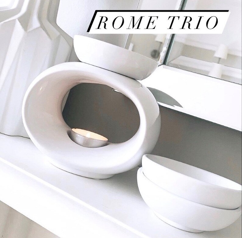 May include: White ceramic wax melt burner with a round opening and a small white candle inside. The burner is on a white surface with two white bowls stacked on top of each other.