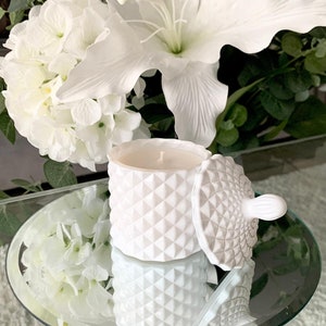 Puede incluir: Vela blanca en un tarro de cer&aacute;mica blanca con un patr&oacute;n geom&eacute;trico. El tarro tiene una tapa con un pomo. La vela est&aacute; sobre una superficie de espejo.