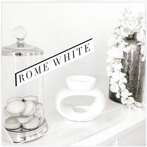 May include: A white ceramic oil burner with a rounded shape and a hole in the centre. The burner is on a white surface with a glass jar filled with tea lights and a silver lid. The text "ROME WHITE" is written in black on a white background.