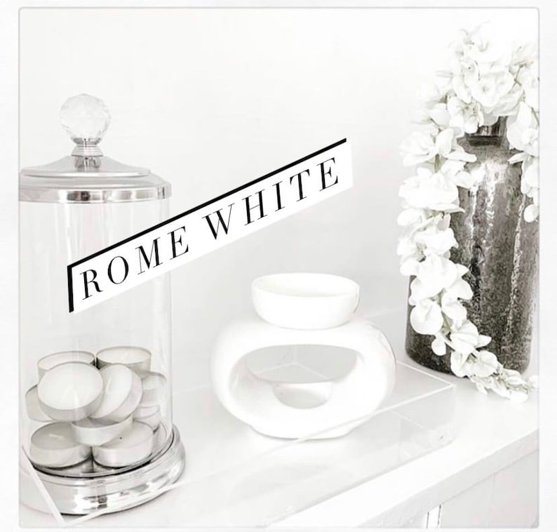May include: A white ceramic oil burner with a rounded shape and a hole in the centre. The burner is on a white surface with a glass jar filled with tea lights and a silver lid. The text "ROME WHITE" is written in black on a white background.