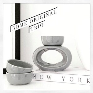 May include: Grey ceramic oil diffuser with a round bowl on top and a larger oval bowl with a hole in the centre. The diffuser is sitting on a book with the words "NEW YORK" printed on the cover. The text "ROME ORIGINAL TRIO" is written in black on a white background.