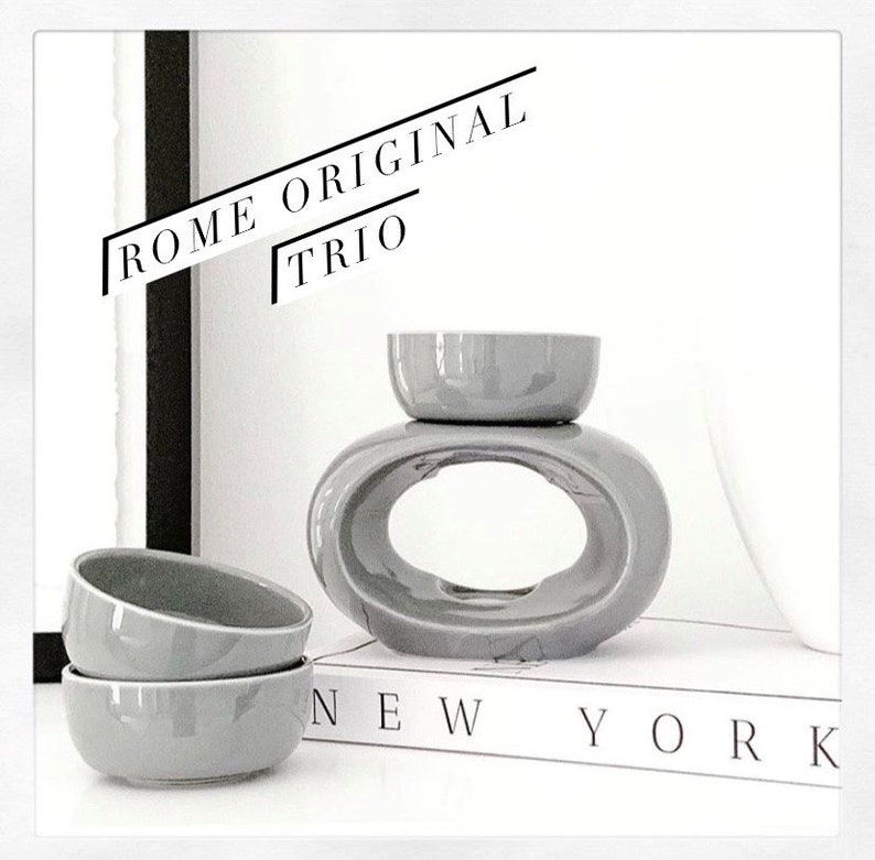 May include: Grey ceramic oil diffuser with a round bowl on top and a larger oval bowl with a hole in the centre. The diffuser is sitting on a book with the words "NEW YORK" printed on the cover. The text "ROME ORIGINAL TRIO" is written in black on a white background.