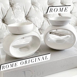 May include: Two white ceramic wax warmers with oval openings. The warmer on the left is labelled "ROME ORIGINAL". The warmer on the right is labelled "ROME".