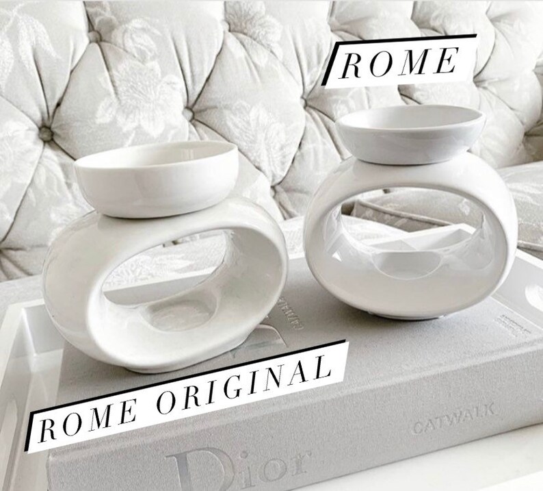 May include: Two white ceramic wax warmers with oval openings. The warmer on the left is labelled "ROME ORIGINAL". The warmer on the right is labelled "ROME".