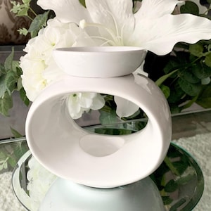 May include: White ceramic wax melt burner with a round, open design. The burner has a small, round dish on top for wax melts.