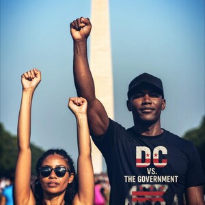 May include: Two people with raised fists in front of the Washington Monument. One wears a black shirt with "DC vs. The Government" and "We Decide Our Future." The other wears a black tank top with "HOME" and a house graphic.