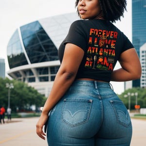Op de afbeelding: Een vrouw draagt een zwart t-shirt met de tekst "FOREVER I LOVE ATLANTA AT NA" in rood, groen en geel. Ze draagt ook een blauwe spijkerbroek. De achtergrond toont een modern gebouw en een stadsstraat.