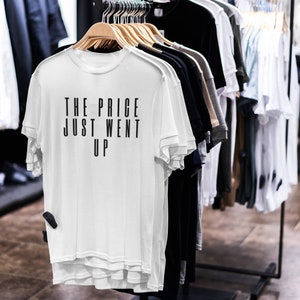 May include: A white t-shirt with the text "THE PRICE JUST WENT UP" in black, hanging on a wooden hanger. The shirt is part of a clothing display with other shirts in various colors, including black, gray, and white. The store setting is visible in the background.