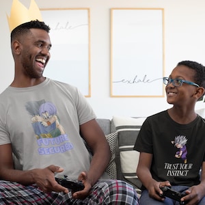 May include: Two people are seated on a gray couch, each holding a game controller. One wears a gray t-shirt with a cartoon graphic and the text "FUTURE SECURED." The other wears a black t-shirt with a graphic and the text "TRUST YOUR INSTINCT."