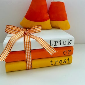May include: A stack of three books painted in orange, yellow, and white with the words "trick or treat" written on them. The books are tied together with a white and orange checkered ribbon and topped with two orange, yellow, and white candy corn pillows.