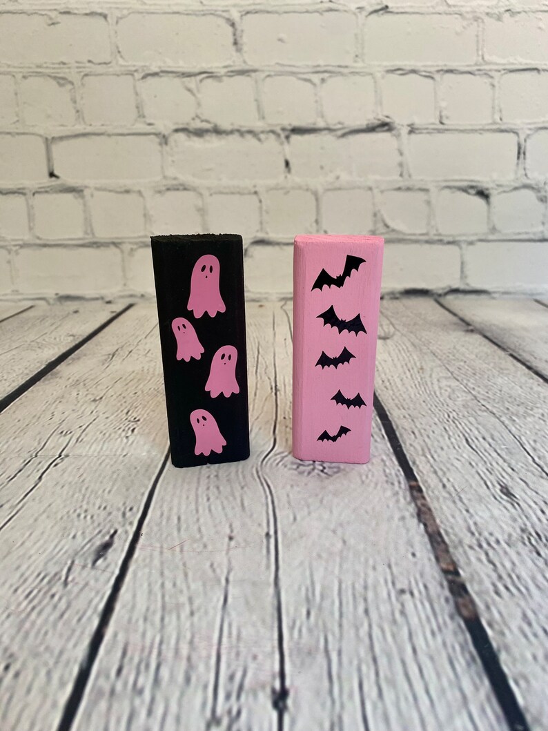 Op de afbeelding: Twee houten blokken, een zwart met roze geesten en een roze met zwarte vleermuizen. Halloween-decoraties.