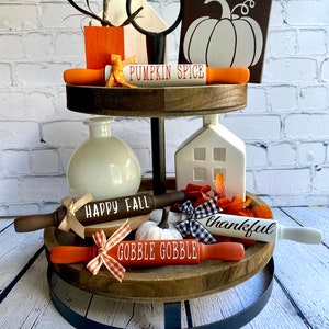 May include: A two-tiered wooden tiered tray with a black metal stand. The tray is decorated with fall-themed items, including a white pumpkin, a white house, a vase, and rolling pins with the words "Happy Fall", "Gobble Gobble", "Thankful", and "Pumpkin Spice".