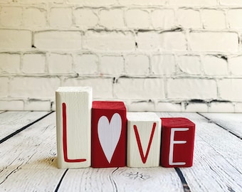 May include: Wooden blocks spelling "LOVE". The "L" and "V" are on white blocks with red lettering. The "O" is a red block with a white heart. The "E" is on a red block with white lettering. The blocks are on a white wooden surface.