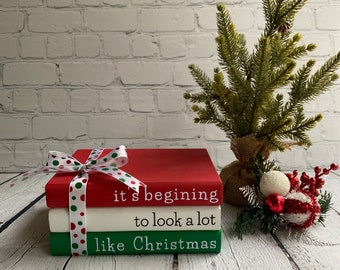 May include: Three stacked books with a red, white, and green color scheme. The top book is red and says "it's beginning to look a lot like Christmas". The books are tied together with a red and green polka dot ribbon.