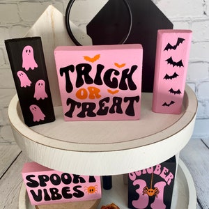 Op de afbeelding: Een wit etagerekastje met twee verdiepingen, versierd met roze en zwarte Halloween-houten blokken. De bovenste verdieping heeft een zwart blok met geestsilhouetten, een roze blok met de tekst "Trick or Treat" in zwarte en oranje letters, en een roze blok met vleermuissilhouetten. De onderste verdieping heeft een roze blok met de tekst "Spooky Vibes" in zwarte letters en een roze blok met de tekst "October 31" in zwarte letters. Beide blokken op de onderste verdieping hebben een kleine oranje spin erop.