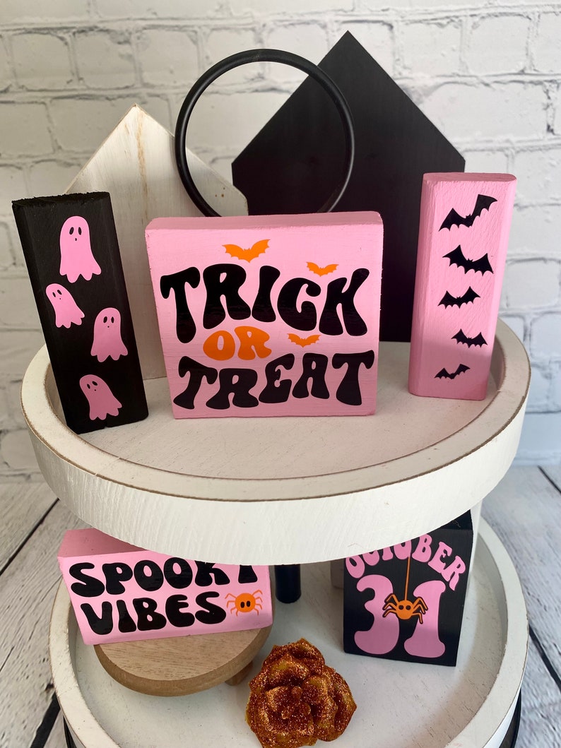 Op de afbeelding: Een wit etagerekastje met twee verdiepingen, versierd met roze en zwarte Halloween-houten blokken. De bovenste verdieping heeft een zwart blok met geestsilhouetten, een roze blok met de tekst "Trick or Treat" in zwarte en oranje letters, en een roze blok met vleermuissilhouetten. De onderste verdieping heeft een roze blok met de tekst "Spooky Vibes" in zwarte letters en een roze blok met de tekst "October 31" in zwarte letters. Beide blokken op de onderste verdieping hebben een kleine oranje spin erop.