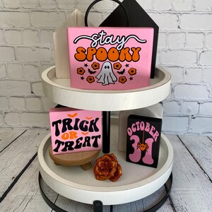 Puede incluir: Una bandeja de dos niveles de color blanco con detalles en metal negro. El nivel superior presenta un letrero de madera rosa con el texto "stay spooky" en letras naranja y blancas, un fantasma y murci&eacute;lagos. El nivel inferior presenta un letrero de madera rosa con el texto "trick or treat" en letras naranja y negras, un letrero de madera negro con el texto "october 31" en letras rosas y una rosa dorada con purpurina.