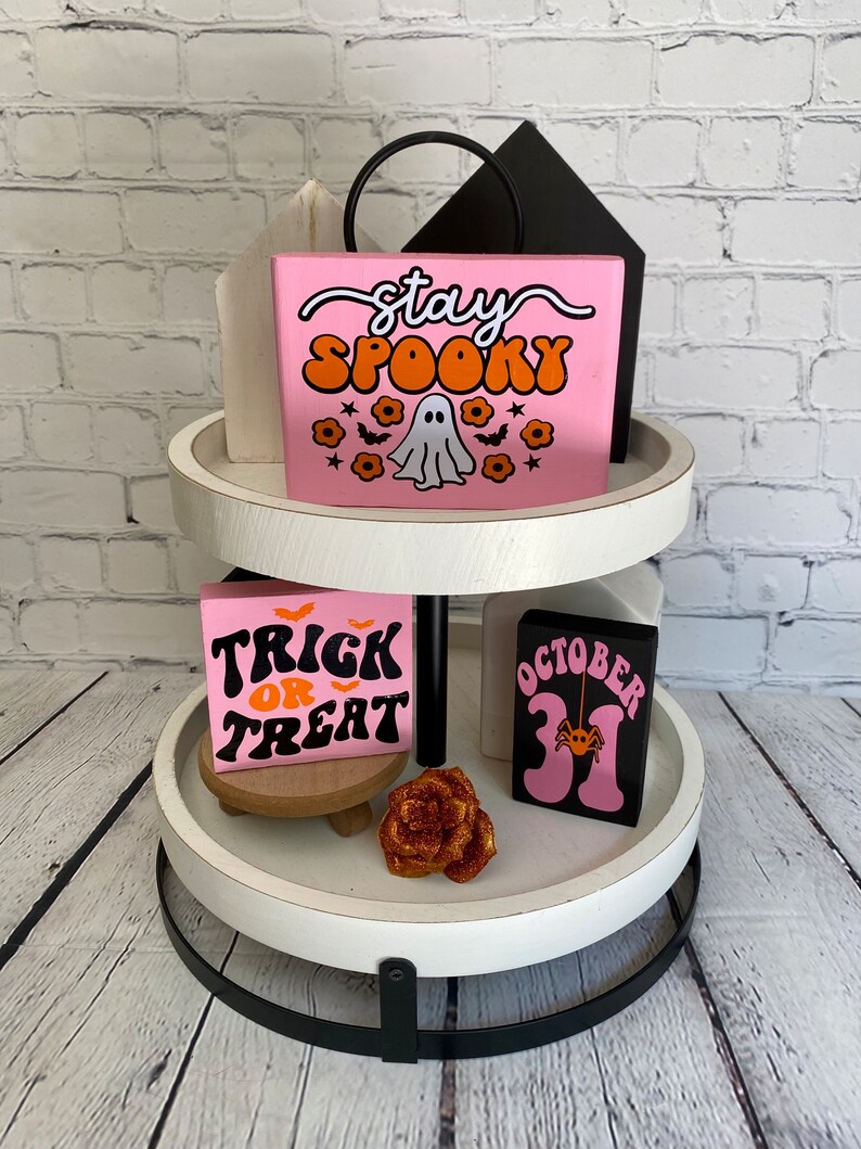 Puede incluir: Una bandeja de dos niveles de color blanco con detalles en metal negro. El nivel superior presenta un letrero de madera rosa con el texto "stay spooky" en letras naranja y blancas, un fantasma y murci&eacute;lagos. El nivel inferior presenta un letrero de madera rosa con el texto "trick or treat" en letras naranja y negras, un letrero de madera negro con el texto "october 31" en letras rosas y una rosa dorada con purpurina.