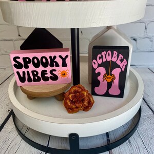 Op de afbeelding: Een wit etag&egrave;re met twee niveaus met een zwarte metalen basis. De bovenste laag heeft drie roze en zwarte houten blokken met de woorden "Trick or Treat" en een ontwerp met een spook en een vleermuis. De onderste laag heeft twee roze en zwarte houten blokken met de woorden "Spooky Vibes" en "October 31" met een spinnenontwerp. Er is een kleine houten standaard met een roze en oranje roos erop.