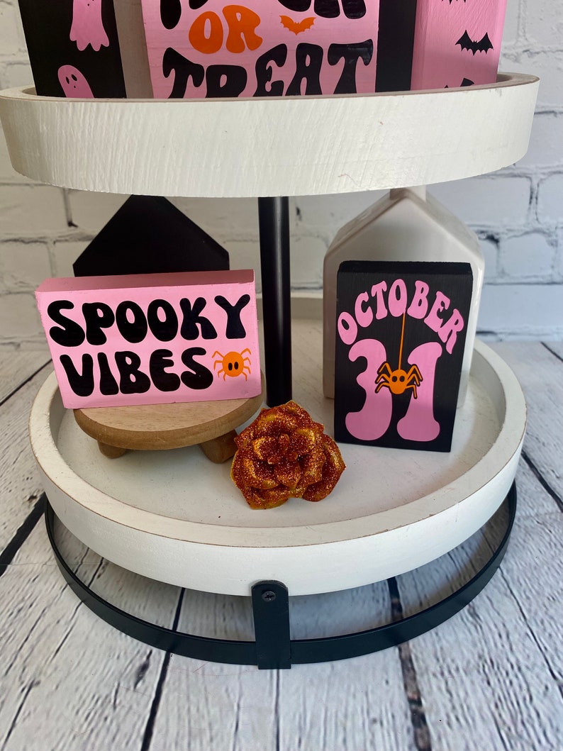 Op de afbeelding: Een wit etag&egrave;re met twee niveaus met een zwarte metalen basis. De bovenste laag heeft drie roze en zwarte houten blokken met de woorden "Trick or Treat" en een ontwerp met een spook en een vleermuis. De onderste laag heeft twee roze en zwarte houten blokken met de woorden "Spooky Vibes" en "October 31" met een spinnenontwerp. Er is een kleine houten standaard met een roze en oranje roos erop.