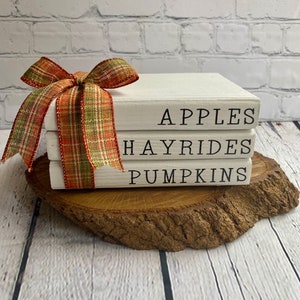 May include: Three white books stacked on a wooden slice. The books are decorated with black lettering that reads "Apples", "Hayrides", and "Pumpkins". A plaid ribbon is tied around the top book.