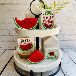 May include: Two-tiered white tiered tray with a black metal base. The tray is decorated with watermelon-themed wooden signs and watermelon slices. The signs read "Hello Summer", "One in a Melon", and "Life is Sweet".