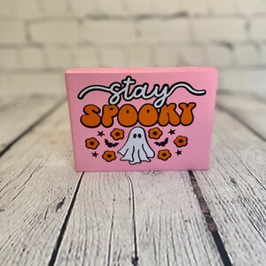 Puede incluir: Bloque de madera rosa con el texto "stay spooky" en letras blancas y naranjas. El bloque presenta un fantasma blanco con una cara negra y flores y murci&eacute;lagos naranjas.