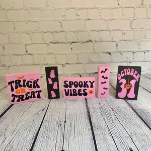 Op de afbeelding: Vijf roze en zwarte houten borden met Halloween-thema's. De borden tonen "Trick or Treat", "Spooky Vibes", "October 31" en hebben geesten en vleermuizen.