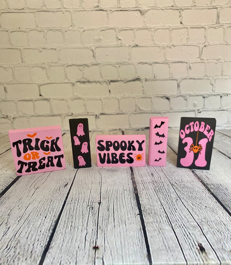 Op de afbeelding: Vijf roze en zwarte houten borden met Halloween-thema's. De borden tonen "Trick or Treat", "Spooky Vibes", "October 31" en hebben geesten en vleermuizen.