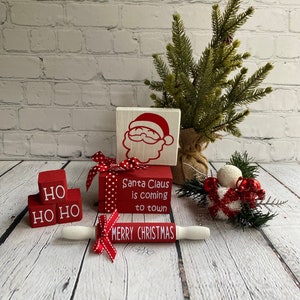 May include: Red and white wooden blocks with the words "HO HO HO" and "Santa Claus is coming to town" on them. A red and white rolling pin with the words "Merry Christmas" on it. A small artificial pine tree with red and white ornaments.