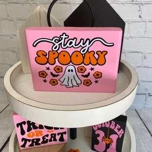 Puede incluir: Un letrero de madera rosa con el texto "stay spooky" en letras blancas y naranjas. Un fantasma blanco con ojos naranjas est&aacute; en el centro del letrero con flores naranjas y murci&eacute;lagos alrededor.
